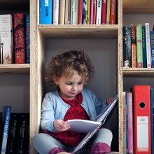 Une petite fille lit un livre assise dans le bas d'une bibliothèque en bois.